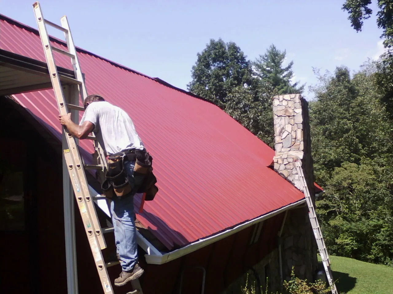 Roofer on a ladder beside a red metal roof and stone chimney — Soffit & Fascia Repair by Roofing Brothers in Swaledale